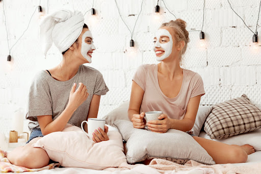 Two smiling young women wearing skin care face masks on a bed while holding mugs.
